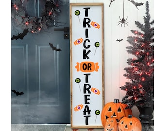 May include: A white wooden sign with the words "Trick or Treat" in black lettering. The sign is decorated with orange and black candy, lollipops, and eyeballs. The sign is leaning against a door with a wreath and bats. There are pumpkins and a Christmas tree in the background.