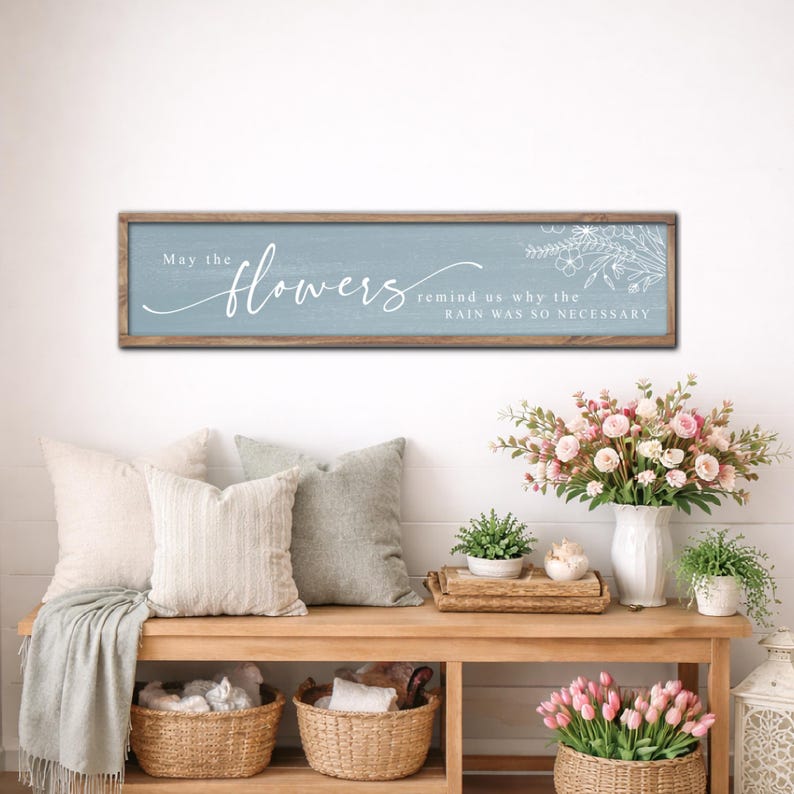 May include: A rectangular wooden sign with the text "May the flowers remind us why the rain was so necessary". The sign is above a wooden bench with cushions, baskets, and floral displays.