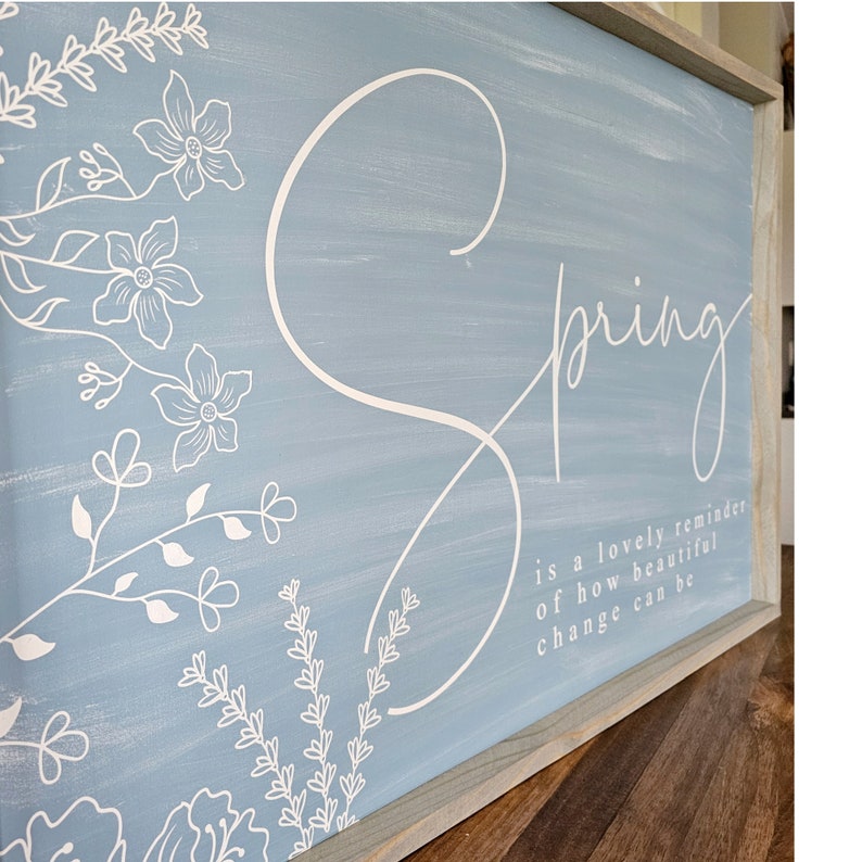 May include: A light blue wooden sign with white floral designs and the word "Spring" in a white cursive font. The sign also includes the text "is a lovely reminder of how beautiful change can be."