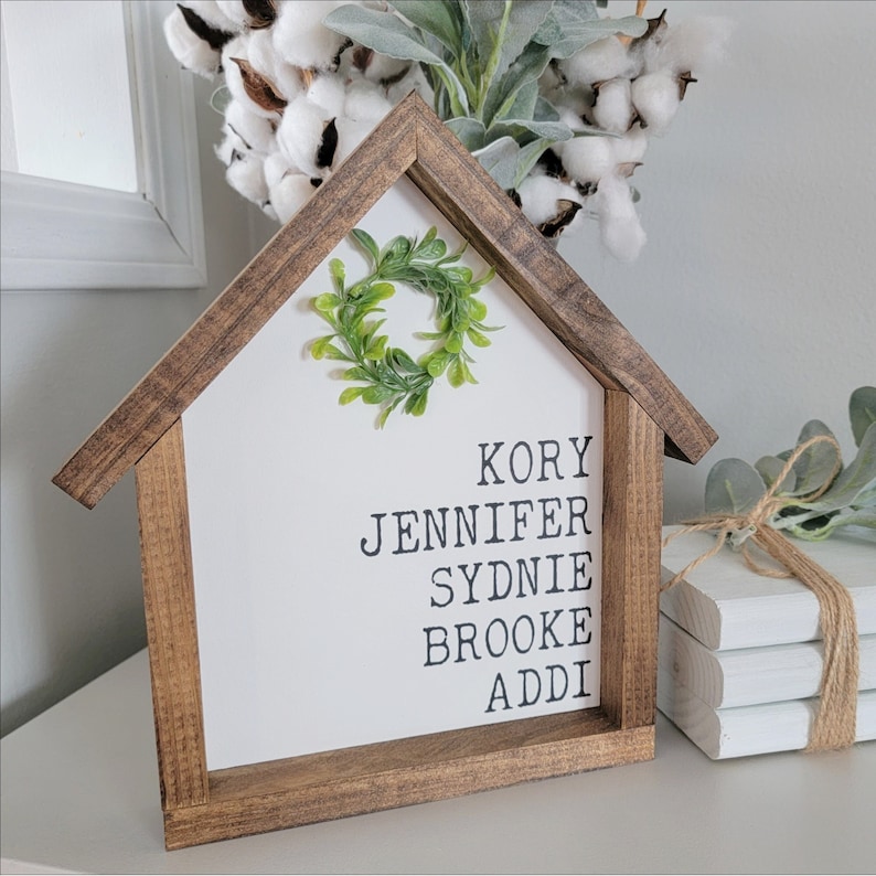 May include: A wooden house-shaped sign with a green wreath on the front. The sign has the names Kory, Jennifer, Sydnie, Brooke, and Addi written on it in black.
