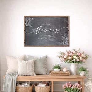 May include: A framed chalkboard sign with the words "May the flowers remind us why the rain was so necessary" in white script. The sign is above a wooden bench with pillows, baskets, and floral arrangements.