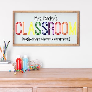 K&ouml;nnte beinhalten: Ein Holzschild mit dem Text "Mrs. Becker's CLASSROOM" in Regenbogenfarben. Unter dem Wort "CLASSROOM" stehen die W&ouml;rter "laugh, share, dream, learn, read". Das Schild steht auf einem Holztisch mit bunten Gegenst&auml;nden.
