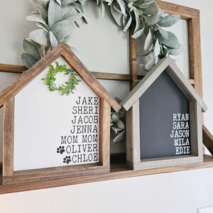 May include: Two wooden house-shaped signs with names listed on them. The sign on the left is white with a green wreath and the names Jake, Sheri, Jacob, Jenna, Mom Mom, Oliver, and Chloe. The sign on the right is black with the names Ryan, Sara, Jason, Mila, and Edie.