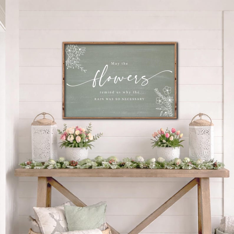 May include: A framed sign with the text "May the flowers remind us why the rain was so necessary" in white script. The sign is on a green background with floral accents. The scene includes a wooden table with floral arrangements and decorative lanterns.