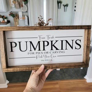 May include: A rectangular wooden sign with the words "PUMPKINS" in large black letters. The sign also includes the phrases "Farm Fresh," "For Pies or Carving," and "Pick Your Own." The sign is held by a person.