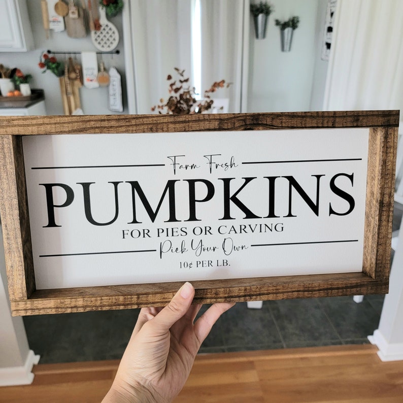 May include: A rectangular wooden sign with the words "PUMPKINS" in large black letters. The sign also includes the phrases "Farm Fresh," "For Pies or Carving," and "Pick Your Own." The sign is held by a person.
