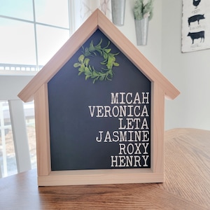 May include: A wooden house-shaped sign with a chalkboard insert. The chalkboard has the names Micah, Veronica, Leta, Jasmine, Roxy, and Henry written in white. A small green wreath is attached to the top of the sign.