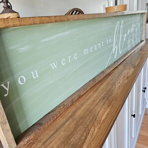 May include: A wooden sign with a light green background and white lettering that reads "You were meant to bloom". The sign is framed in a natural wood frame and has a distressed look.