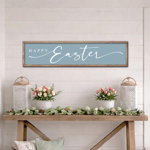 May include: A rectangular wooden sign with a light blue background and the words "HAPPY Easter" in white script. The sign is mounted above a wooden console table decorated with flowers, lanterns, and greenery, creating a festive Easter display.