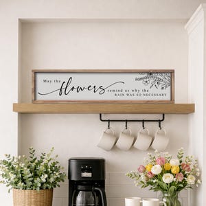 May include: A wooden sign with the text "May the flowers remind us why the rain was so necessary". The sign has a light grey background and floral illustrations. It is displayed above a shelf, with a black mug rack below.