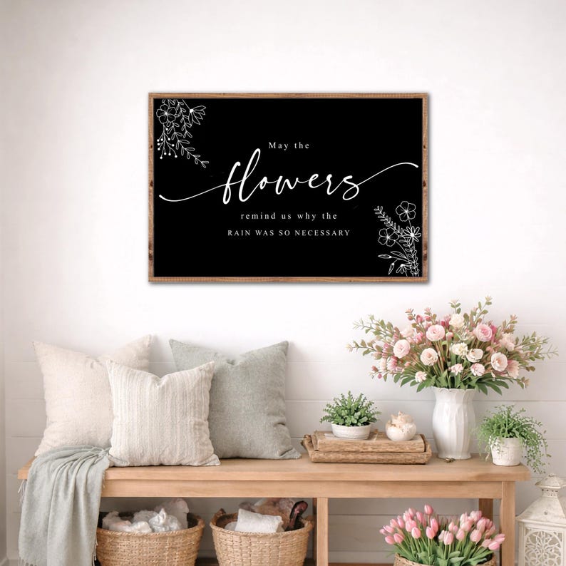 May include: A framed black sign with white script that reads "May the flowers remind us why the rain was so necessary." Floral illustrations are in the corners. The sign is above a wooden bench with pillows, baskets, and flowers.