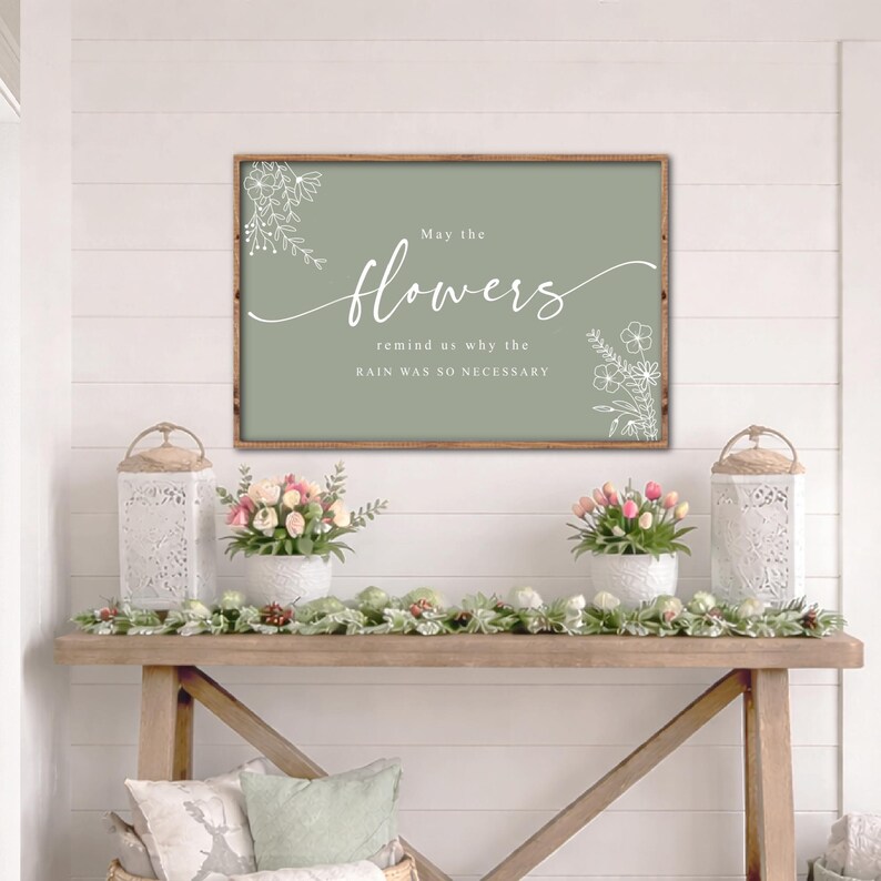 May include: A framed sign with the text "May the flowers remind us why the rain was so necessary" in white script on a sage green background. The sign is above a wooden console table with floral arrangements and decorative lanterns.