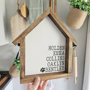 May include: A wooden house-shaped sign with the names Holden, Emma, Collins, Oaklyn, and Bentley written in black lettering. A paw print is below the names.