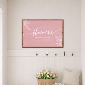 May include: A framed pink sign with the words "flowers" in cursive and the phrase "May the flowers remind us why the rain was so necessary." White floral illustrations adorn the corners. The frame is a light brown color.