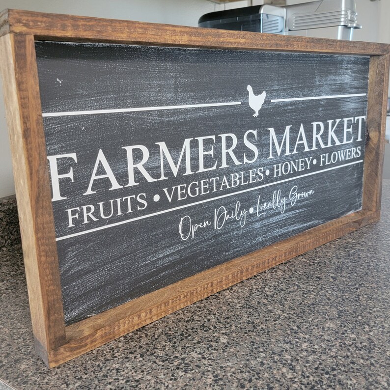 K&ouml;nnte beinhalten: Ein rustikales Schild mit Holzrahmen, schwarzem Hintergrund aus Schiefertafel und wei&szlig;em Text, der "Farmers Market Fruits Vegetables Honey Flowers Open Daily Locally Grown" lautet, mit einer wei&szlig;en Silhouette eines Huhns &uuml;ber dem Wort "Market".
