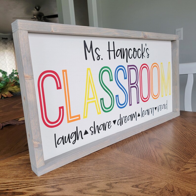 K&ouml;nnte beinhalten: Rechteckiges Holzschild mit grauem Rahmen. Auf dem Schild steht "Ms. Hancock's CLASSROOM" in bunten Buchstaben. Unter dem Wort "CLASSROOM" stehen die W&ouml;rter "laugh, share, dream, learn, read".