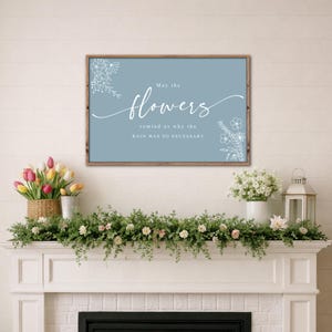 May include: A framed sign with the text "May the flowers remind us why the rain was so necessary" in white script on a blue background. The sign is surrounded by a brown wooden frame. The image also shows a fireplace mantel decorated with flowers and greenery.