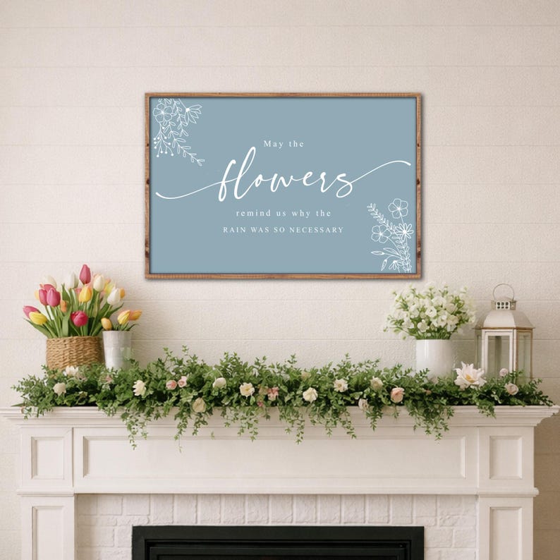 May include: A framed sign with the text "May the flowers remind us why the rain was so necessary" in white script on a blue background. The sign is surrounded by a brown wooden frame. The image also shows a fireplace mantel decorated with flowers and greenery.