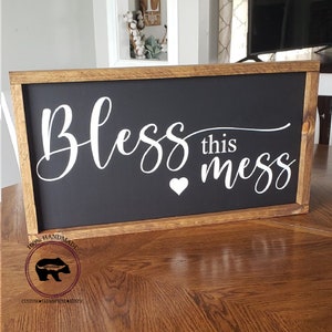 May include: A black framed wooden sign with white lettering that reads "Bless this mess" with a small white heart. The sign is handmade and has a rustic look.