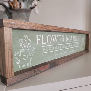 以下が含まれることがあります： 緑色の背景に白い文字で「FLOWER MARKET - BLOOMS - SEEDS - STEMS - Open every Saturday & Sunday」と書かれた木製の看板。看板には、花が入ったジョウロの白い輪郭が描かれている。