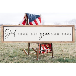 May include: A wooden sign with the text "God shed his grace on thee" in black cursive lettering. The sign is hanging on a rocking chair with an American flag draped over it.