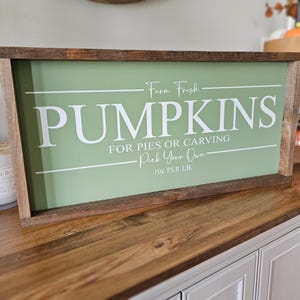 May include: A rectangular wooden sign with a light green background and white text that reads "PUMPKINS FOR PIES OR CARVING Pick Your Own". The sign has a dark brown wooden frame. The text "Farm Fresh" is above "PUMPKINS".