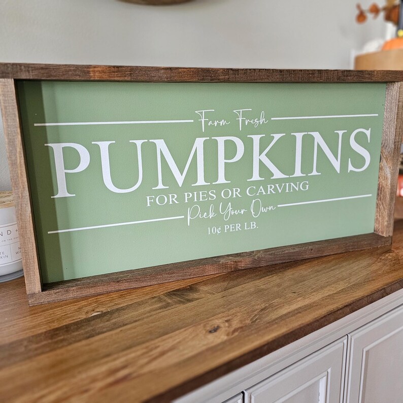 May include: A rectangular wooden sign with a light green background and white text that reads "PUMPKINS FOR PIES OR CARVING Pick Your Own". The sign has a dark brown wooden frame. The text "Farm Fresh" is above "PUMPKINS".