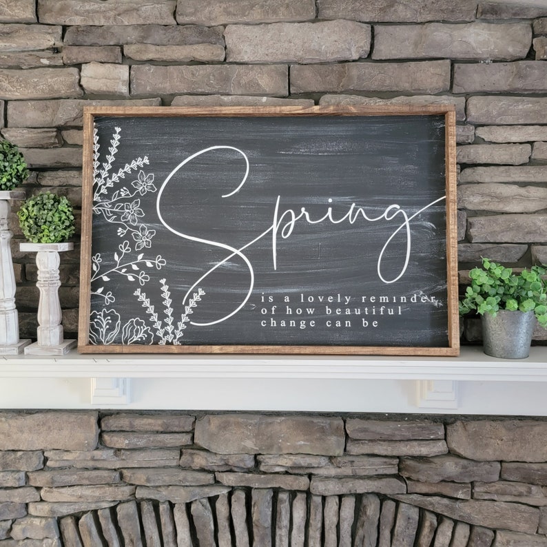 May include: A black chalkboard sign with a white wooden frame. The sign says "Spring is a lovely reminder of how beautiful change can be." The word "Spring" is in a large, cursive font and is surrounded by white floral designs.