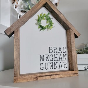 May include: A wooden house-shaped sign with a green wreath on top. The sign has the names "Brad, Meghan, Gunnar" written in black lettering.