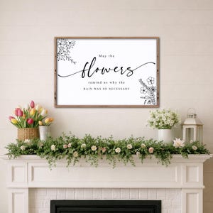 May include: A framed wall art print with the text "May the flowers remind us why the rain was so necessary." The print has a white background with black floral illustrations and text. The frame is brown.