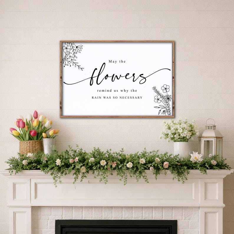 May include: A framed wall art print with the text "May the flowers remind us why the rain was so necessary." The print has a white background with black floral illustrations and text. The frame is brown.
