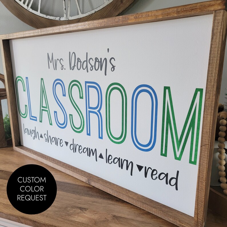 K&ouml;nnte beinhalten: Ein rechteckiges, holzgerahmtes Schild mit den Worten "Mrs. Dodson's CLASSROOM" in gr&uuml;nen und blauen Buchstaben. Unter dem Wort "CLASSROOM" stehen die Worte "laugh, share, dream, learn, read". Ein schwarzer Kreis mit den Worten "CUSTOM COLOR REQUEST" befindet sich in der linken unteren Ecke.