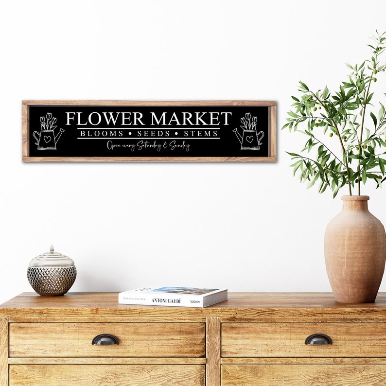 以下が含まれることがあります： 「FLOWER MARKET」と「BLOOMS - SEEDS - STEMS」の文字と、両側にジョウロと花のグラフィックが描かれた白黒の看板。看板には「Open every Saturday & Sunday」とも書かれています。