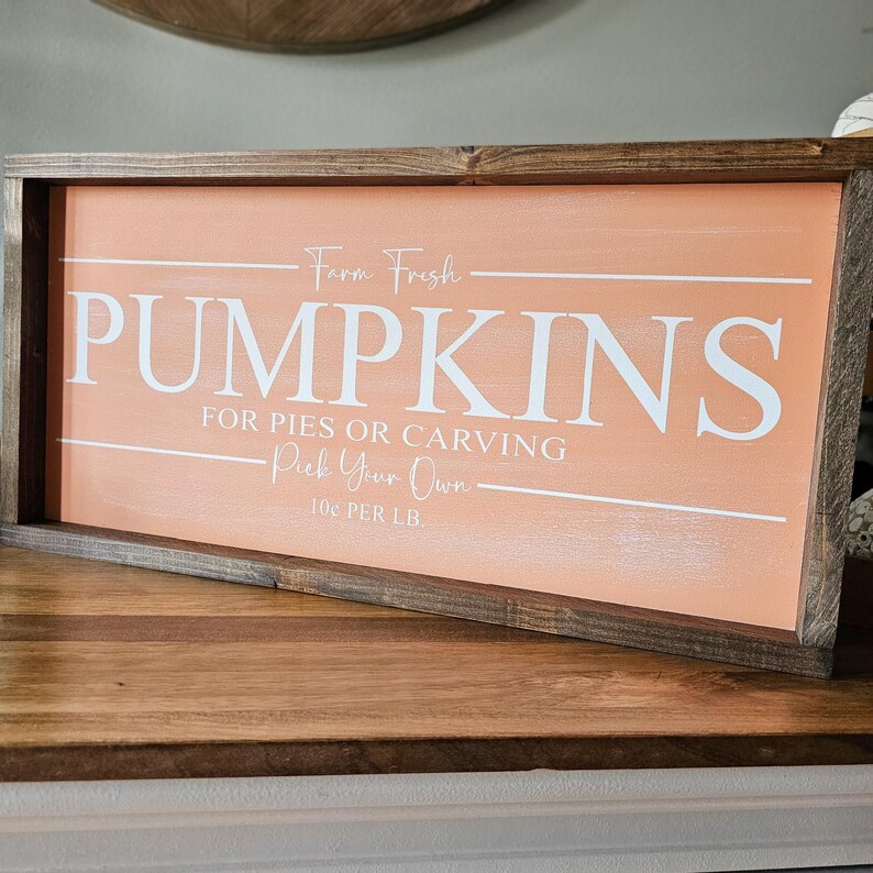 May include: Wooden framed sign with a peach-colored background and white text that reads "PUMPKINS" and "Farm Fresh". The sign also says "For Pies or Carving" and "Pick Your Own".