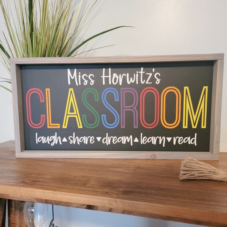 K&ouml;nnte beinhalten: Ein gerahmtes Tafelschild mit dem Text "Miss Horwitz's CLASSROOM" in bunten Buchstaben. Darunter stehen die Worte "laugh, share, dream, learn, read". Der Rahmen ist hellgrau und der Hintergrund ist schwarz.