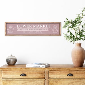 以下が含まれることがあります： ピンクの木製の看板に白い文字で「FLOWER MARKET」と書かれており、その下に「BLOOMS, SEEDS, STEMS」と書いてあります。看板には「Open every Saturday & Sunday」とも書かれており、両側に花が咲いたジョウロの絵が描かれています。