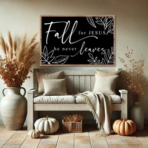 May include: A black and white sign with the text "Fall for Jesus, he never leaves" with white leaf illustrations. The sign is hanging on a wall above a wooden bench with pillows and a throw blanket. There are two pumpkins and a vase with dried flowers on the floor.
