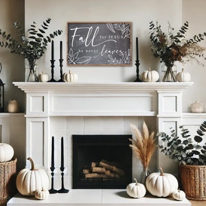 May include: A white farmhouse style fireplace mantel with a black framed sign that reads "Fall for Jesus he never leaves" with white lettering and a decorative leaf design. The mantel is decorated with white pumpkins, greenery, and candlesticks.