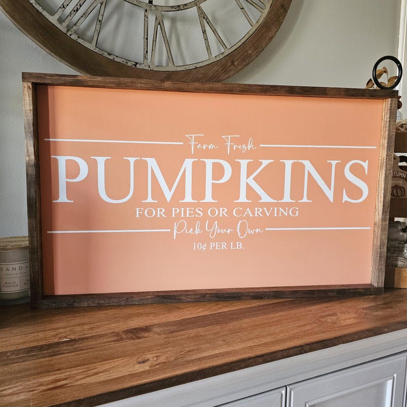 May include: A rectangular wooden sign with a peach-colored background and white text that reads "Farm Fresh PUMPKINS FOR PIES OR CARVING Pick Your Own 10&cent; PER LB." The sign has a dark brown wooden frame.