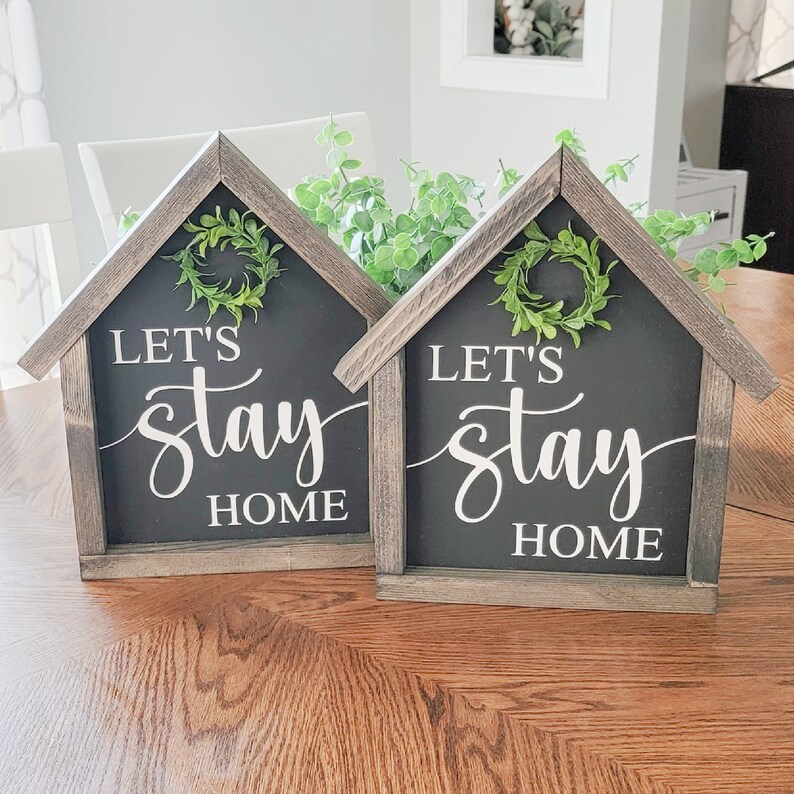 May include: Two wooden signs shaped like houses with black chalkboard backgrounds and white lettering that reads "Let's stay home". Each sign has a small green wreath on the front.
