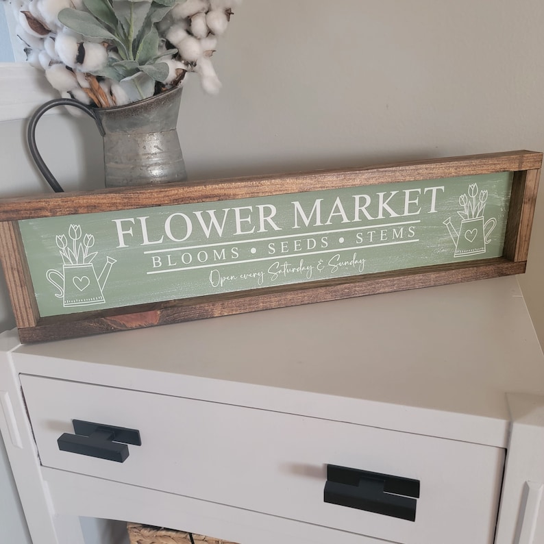 以下が含まれることがあります： 緑色の背景に白い文字で「Flower Market Blooms Seeds Stems Open every Saturday & Sunday」と書かれた木製の看板。看板は茶色の枠で囲まれており、花とハートが描かれた白いじょうろが2つ描かれている。