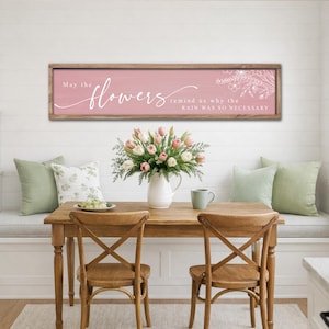 May include: A wooden framed sign with the text "May the flowers remind us why the rain was so necessary" in white script on a pink background. A bouquet of tulips sits on a wooden table with two chairs.