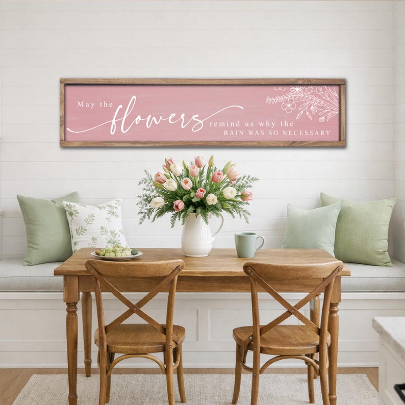 May include: A wooden framed sign with the text "May the flowers remind us why the rain was so necessary" in white script on a pink background. A bouquet of tulips sits on a wooden table with two chairs.