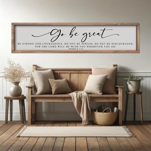 May include: A wooden framed sign with the words "Go be great" in elegant script. Below, the text reads "Be strong and courageous...Joshua 1:9." The sign hangs above a wooden bench with pillows and a woven basket, creating a cozy, inspirational home decor scene.