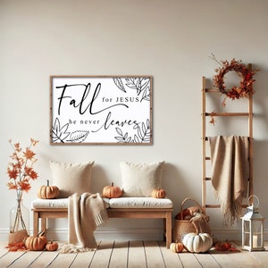 May include: A white sign with black text and leaf illustrations reads "Fall for Jesus he never leaves". The sign is hanging on a white wall above a wooden bench with pillows and a blanket. There are pumpkins and fall decor on the floor.