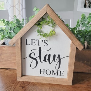 May include: A wooden house-shaped sign with a white background and black lettering that reads "Let's stay home". A small green wreath is attached to the top of the sign.