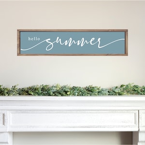 May include: A wooden sign with a light blue background and white lettering that says "hello summer" in a cursive font. The sign is hanging on a white wall above a white mantel with a green garland.