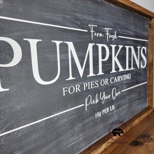 May include: A rectangular wooden sign with a dark gray background and white lettering. The sign reads "Farm Fresh PUMPKINS FOR PIES OR CARVING Pick Your Own 10&cent; PER LB." The sign has a brown wooden frame and a bear logo.