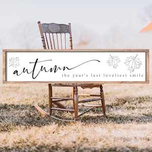 May include: A rectangular wooden sign with a white background and a brown frame. The sign features the word "autumn" in cursive, along with the phrase "the year's last loveliest smile" and leaf illustrations. The sign is displayed outdoors.