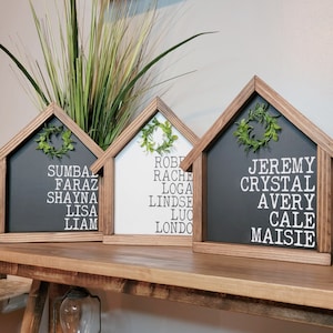 May include: Three wooden house-shaped signs with black, white, and grey backgrounds. Each sign has a wreath and a list of names in white lettering. The names on the signs are: Sumbaz, Faraz, Shayna, Lisa, Liam, Robe, Rache, Loga, Lindse, Luc, Londo, Jeremy, Crystal, Avery, Cale, Maisie.